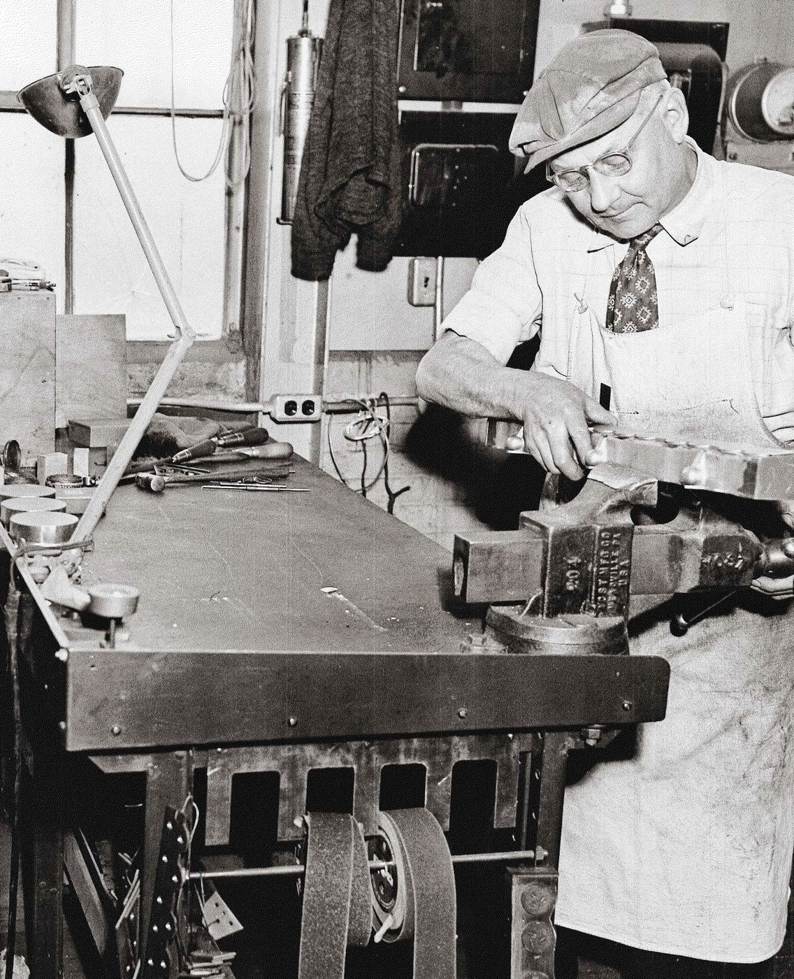 An older Anderson Pattern craftsman in tie and apron working at a vise